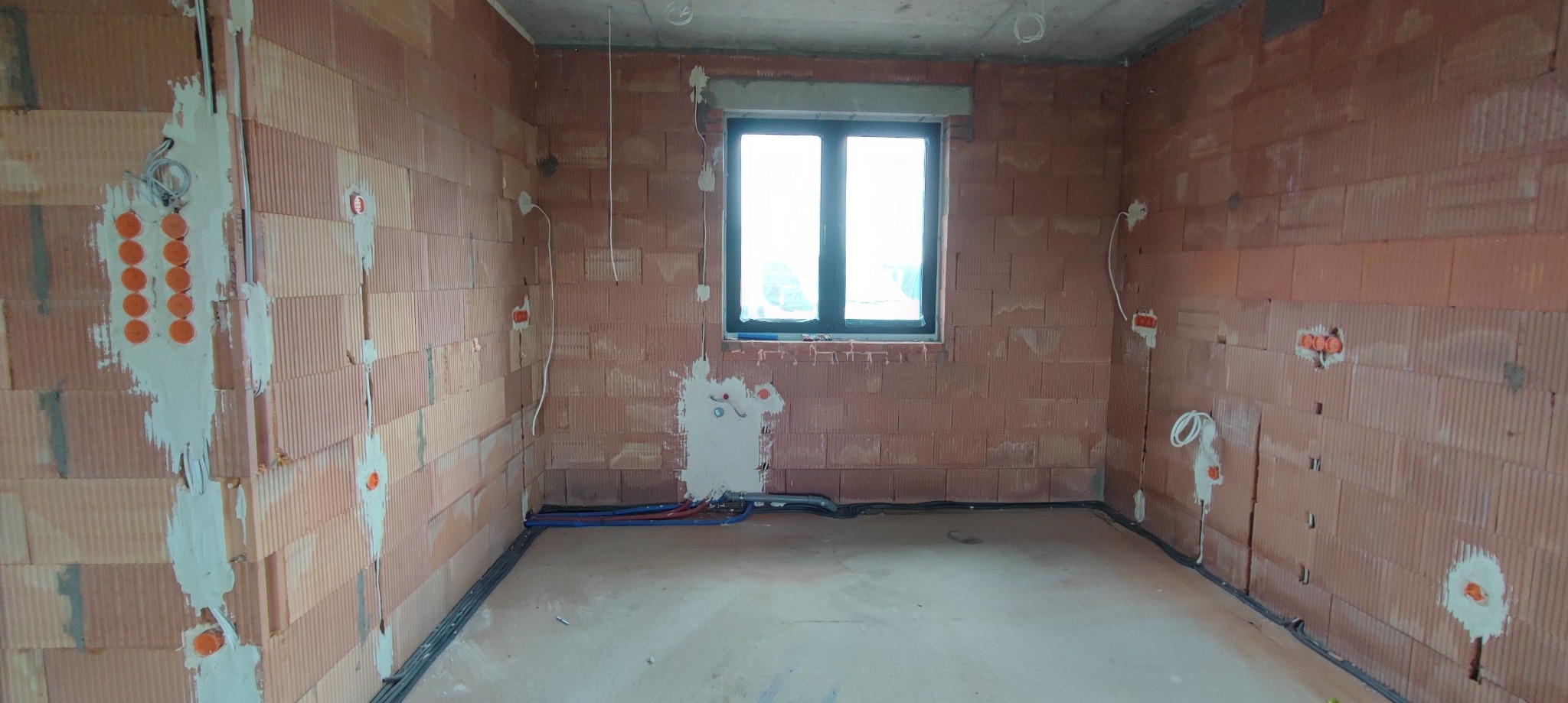 Surowy stan wnętrza domu z widoczną instalacją elektryczną w trakcie montażu, ściany z czerwonej cegły, okno z ciemną ramą, kable i puszki elektryczne wystające ze ścian.