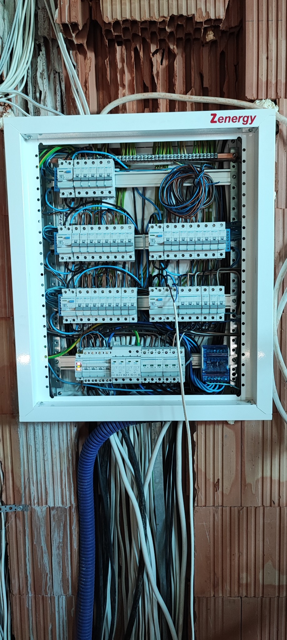 Rozdzielnica elektryczna Zenergy zamontowana na ścianie z pustaków ceramicznych, z widocznymi zabezpieczeniami i przewodami w różnych kolorach, w tym niebieska rura karbowana doprowadzająca kable.