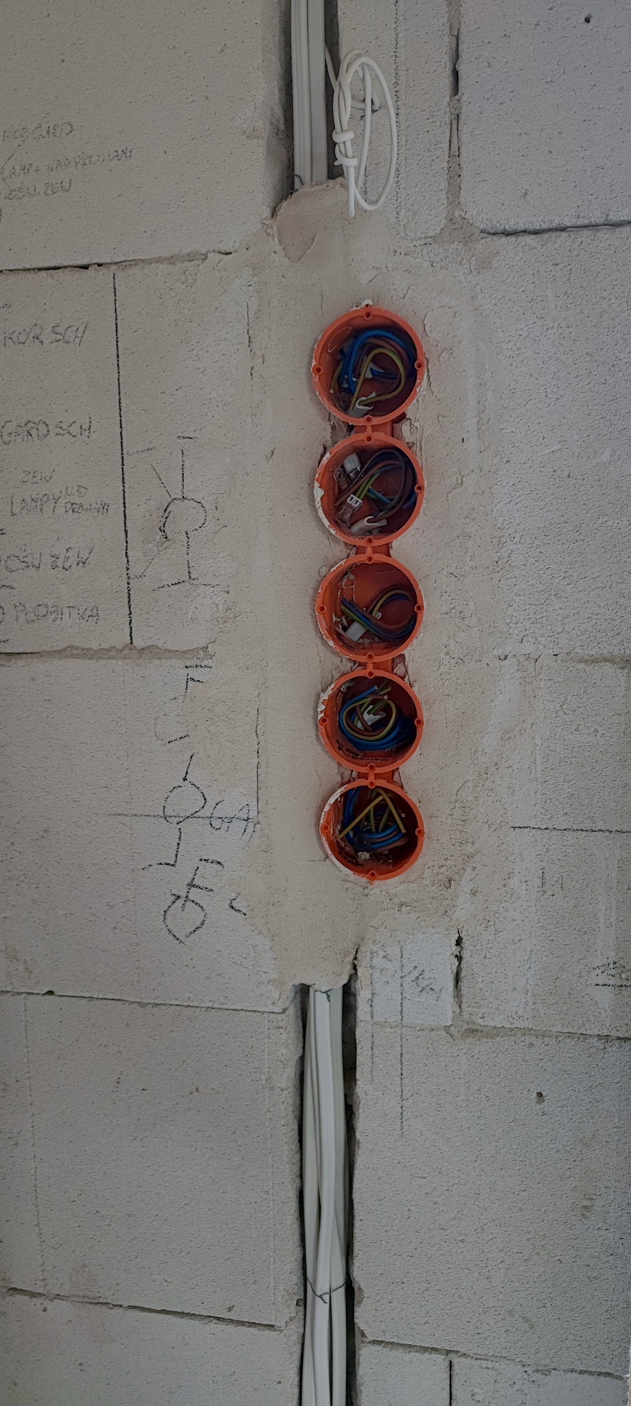 Surowa ściana z betonu komórkowego z czterema pomarańczowymi puszkami instalacyjnymi w pionowym rzędzie, zawierającymi kolorowe przewody elektryczne; obok widoczne pionowe korytka kablowe...
