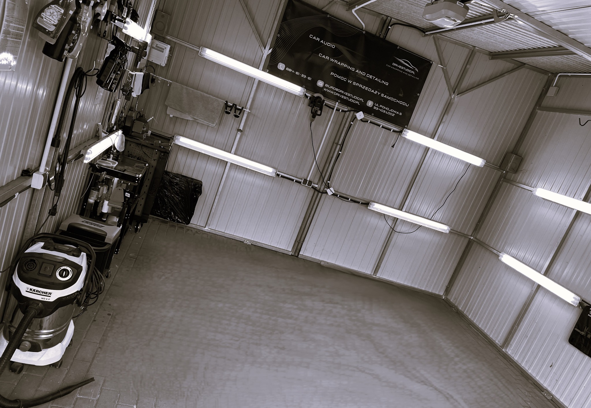 Wnętrze warsztatu samochodowego z widocznym odkurzaczem Karcher WD 6 P, regałem z narzędziami i banerem reklamowym z informacjami kontaktowymi firmy zajmującej się car audio, oklejaniem...