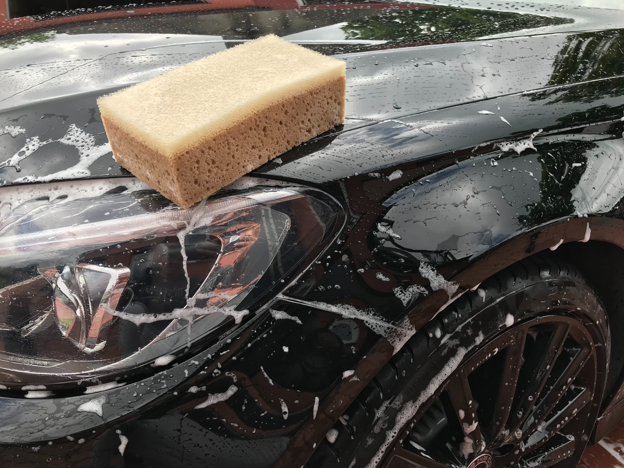 Czarny samochód z pianą i gąbką na karoserii, widoczny fragment opony i reflektor.