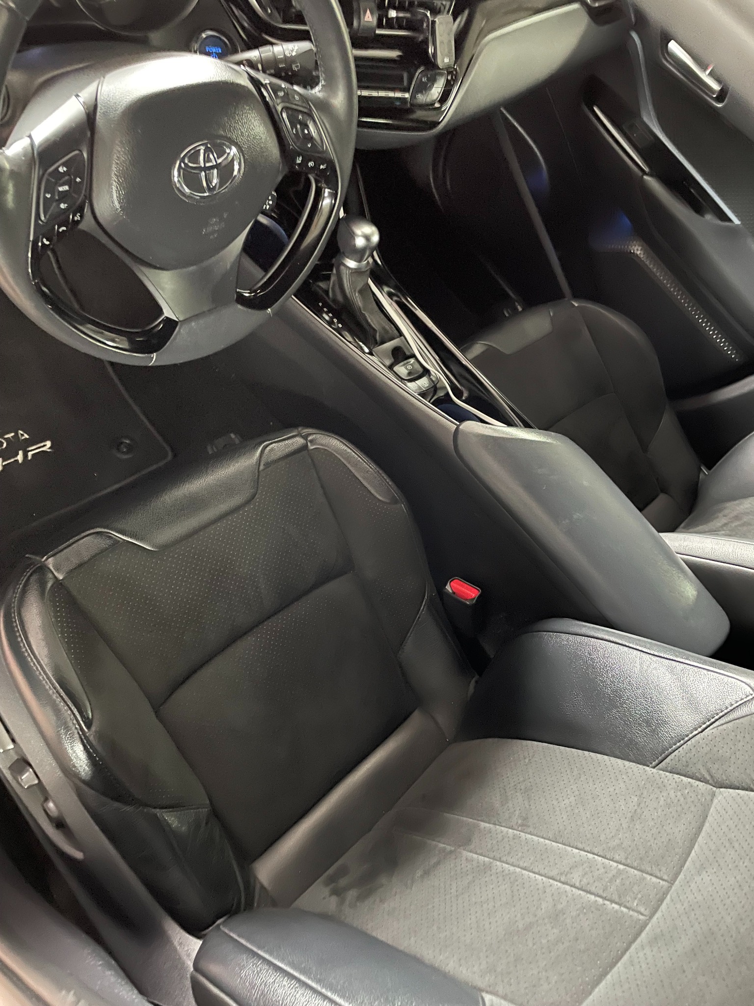 Wnętrze samochodu Toyota C-HR z czarną skórzaną tapicerką, widoczne fotele, kierownica z logo Toyota oraz konsola środkowa. Na podłodze widoczny dywanik z napisem C-HR.