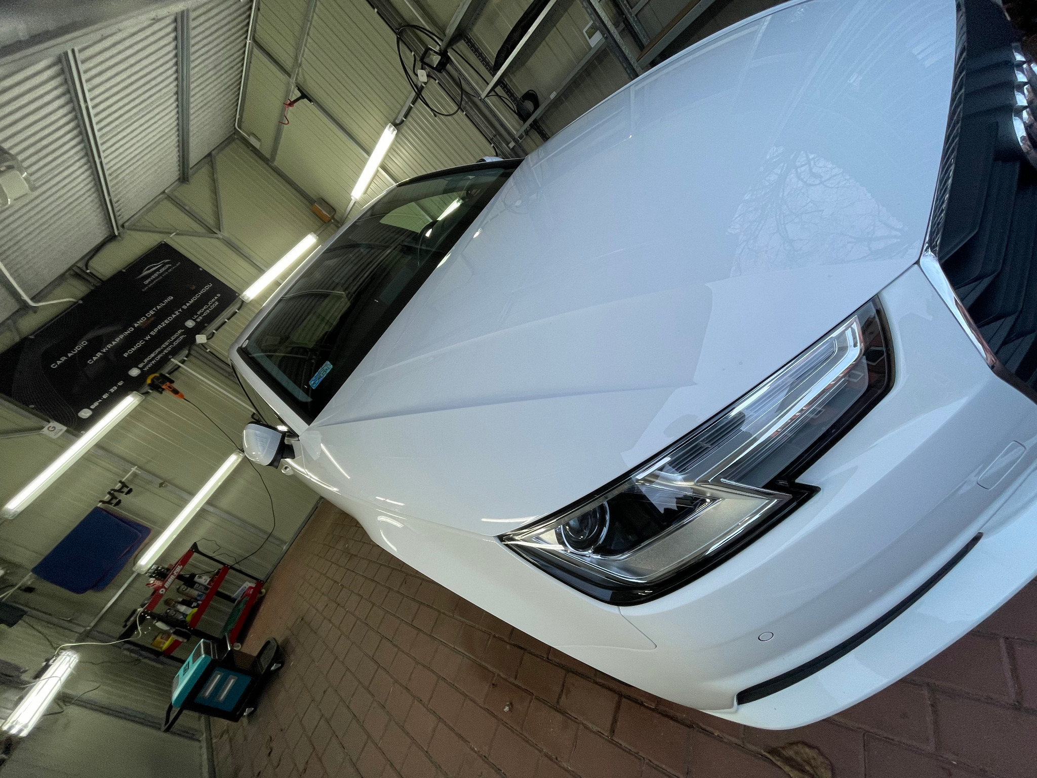 Biały samochód Audi po umyciu w warsztacie, widoczny reflektor, błyszcząca karoseria, na podłodze ceglana kostka, w tle regał z preparatami i sprzęt do czyszczenia.