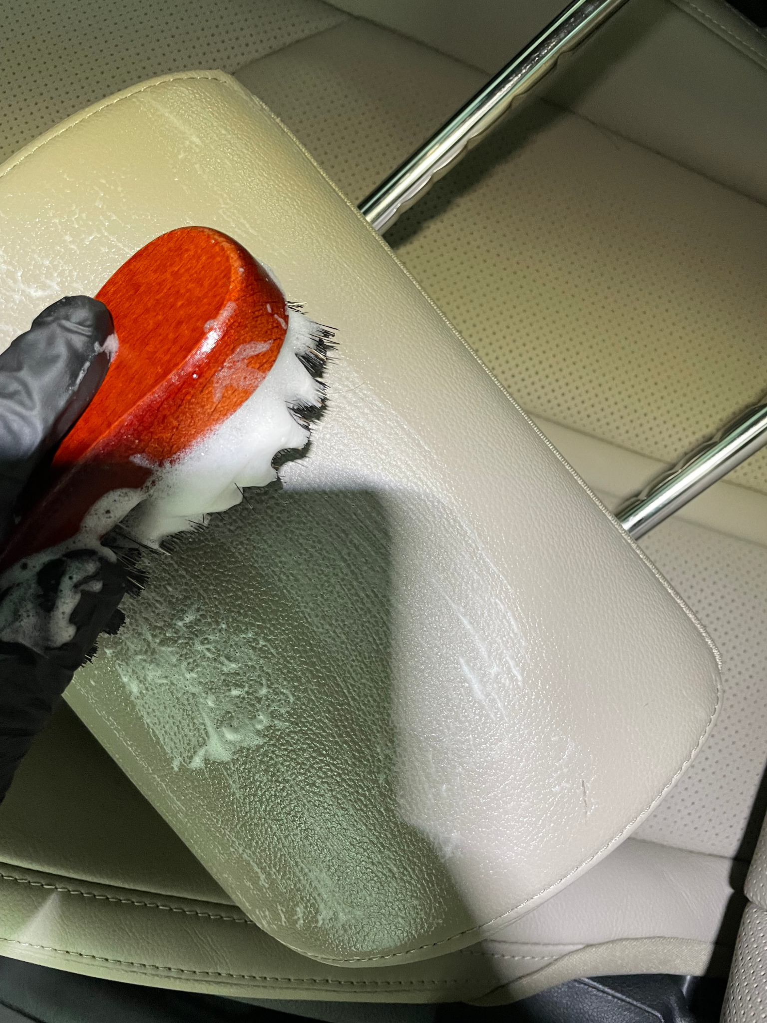 Szczotka z drewnianą rączką i białym detergentem w czarnej rękawicy czyści zagłówek samochodowy z jasnej skóry.