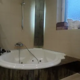 Biała, narożna wanna z hydromasażem, obudowana panelami imitującymi stare drewno, na tle beżowych płytek i dekoracyjnej kolumny z podświetleniem w nowoczesnej łazience. Na parapecie okna widoczna...