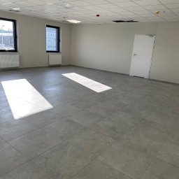 Fast Serwis Karol Majka - Wykończone wnętrze biurowe z szarą podłogą z płytek, białymi ścianami i sufitem kasetonowym. Dwa okna z grzejnikami i białe drzwi po prawej stronie.