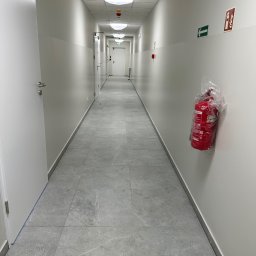 Fast Serwis Karol Majka - Długi korytarz biurowy z szarą podłogą z płytek, białymi ścianami, białymi drzwiami i widocznym znakiem ewakuacyjnym oraz gaśnicą w folii ochronnej.