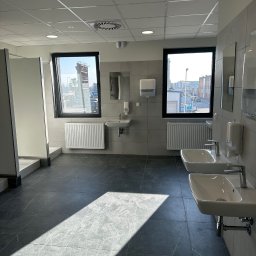 Fast Serwis Karol Majka - Jasne, przestronne wnętrze toalety publicznej z umywalkami, grzejnikami i kabinami. Widoczne płytki podłogowe imitujące marmur i okna z widokiem na zewnątrz.