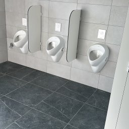 Fast Serwis Karol Majka - Nowoczesna toaleta publiczna z trzema białymi pisuarami i szarymi płytkami na ścianach i podłodze. Minimalistyczny design, czyste linie, oświetlenie górne.