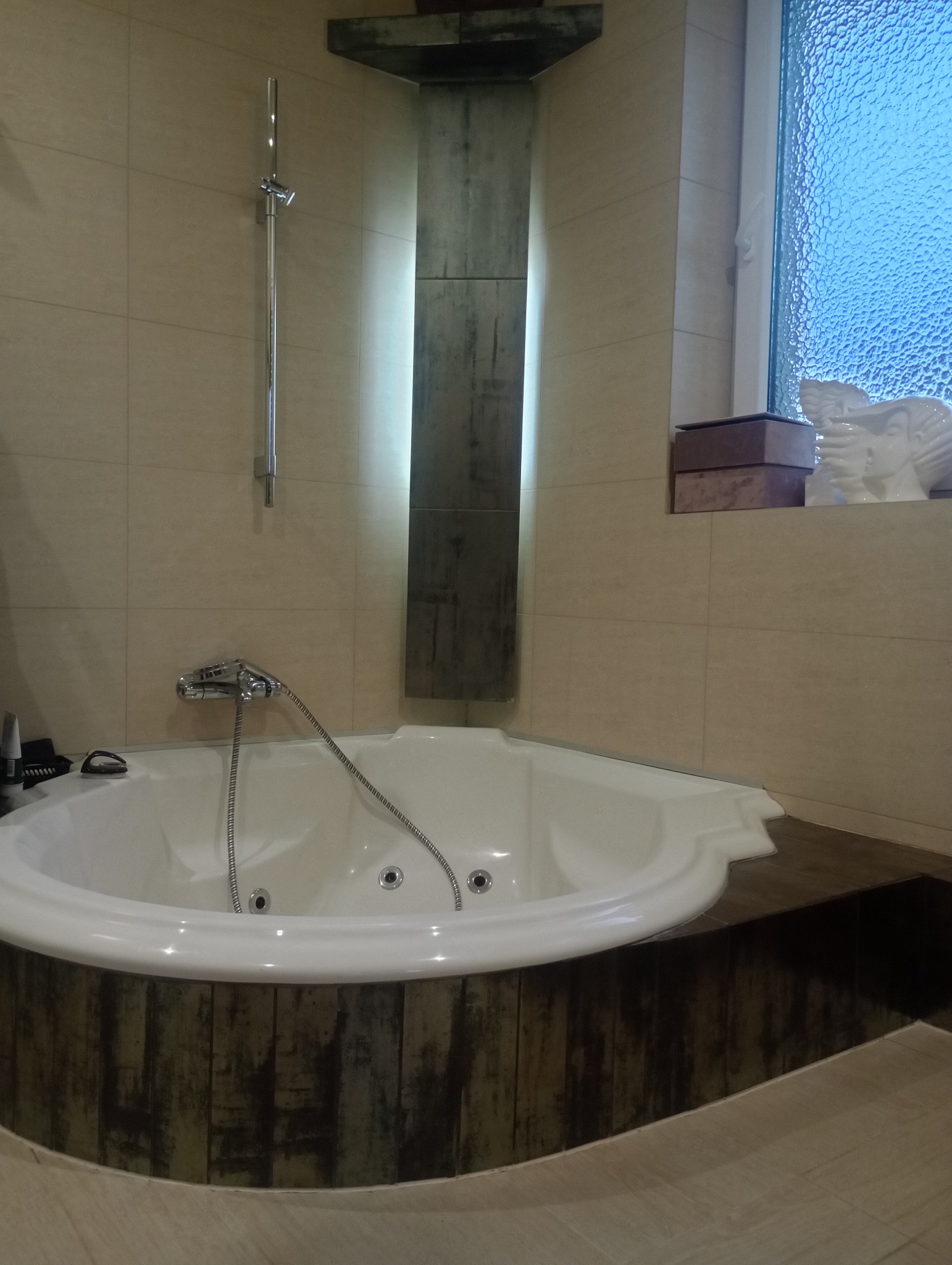 Biała, narożna wanna z hydromasażem, obudowana panelami imitującymi stare drewno, na tle beżowych płytek i dekoracyjnej kolumny z podświetleniem w nowoczesnej łazience. Na parapecie okna widoczna...