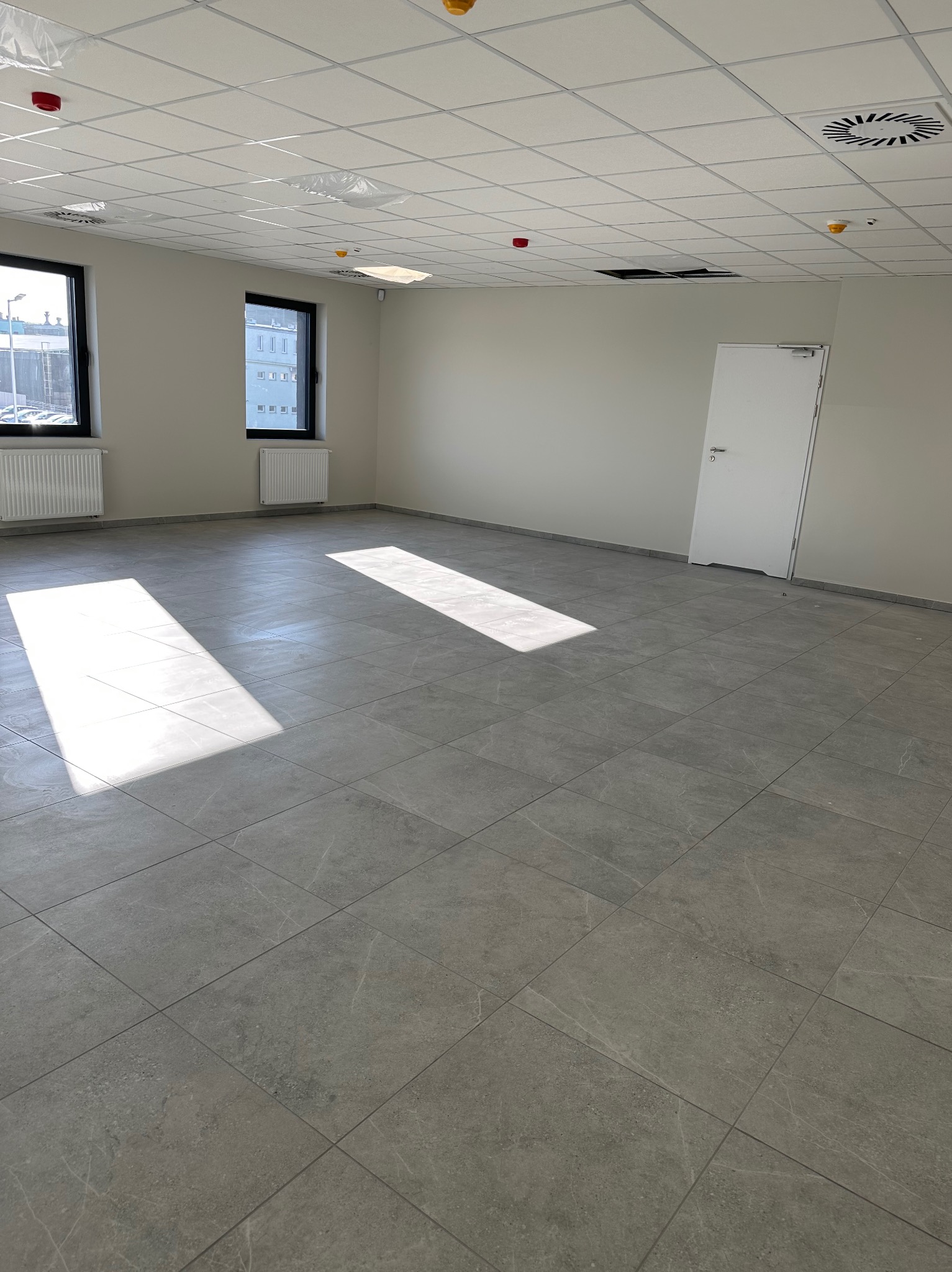 Wykończone wnętrze biurowe z szarą podłogą z płytek, białymi ścianami i sufitem kasetonowym. Dwa okna z grzejnikami i białe drzwi po prawej stronie.
