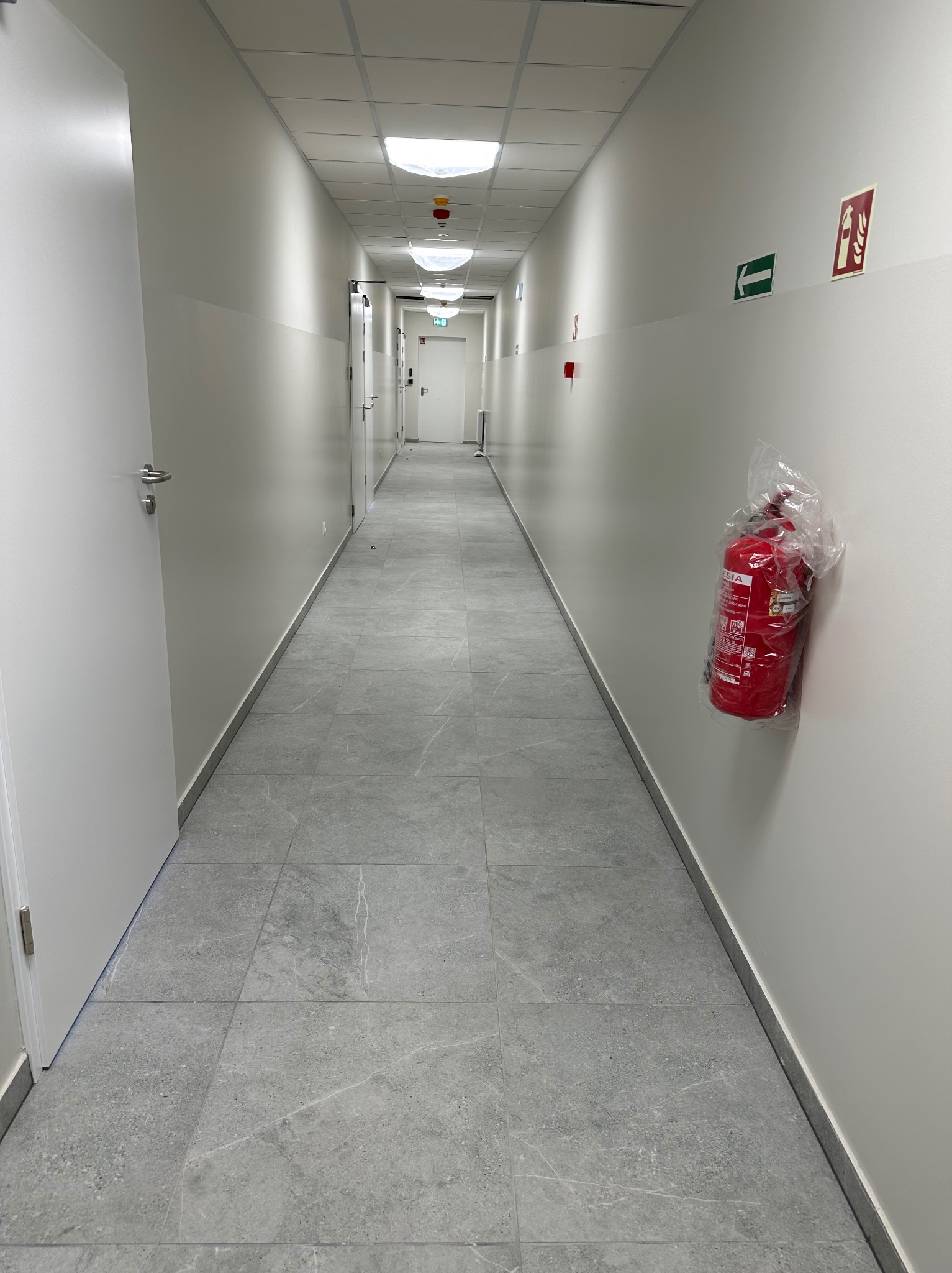 Długi korytarz biurowy z szarą podłogą z płytek, białymi ścianami, białymi drzwiami i widocznym znakiem ewakuacyjnym oraz gaśnicą w folii ochronnej.