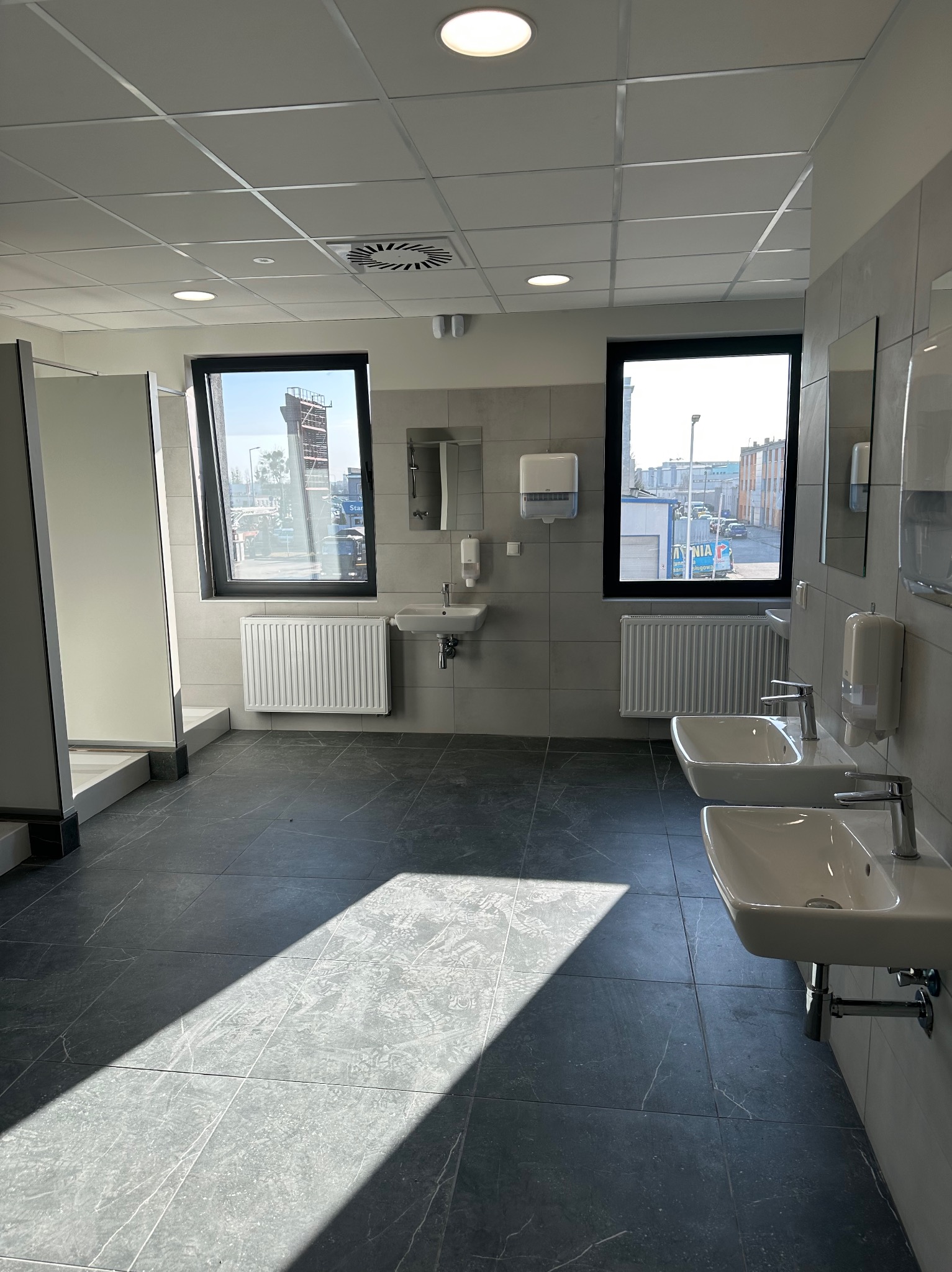 Jasne, przestronne wnętrze toalety publicznej z umywalkami, grzejnikami i kabinami. Widoczne płytki podłogowe imitujące marmur i okna z widokiem na zewnątrz.