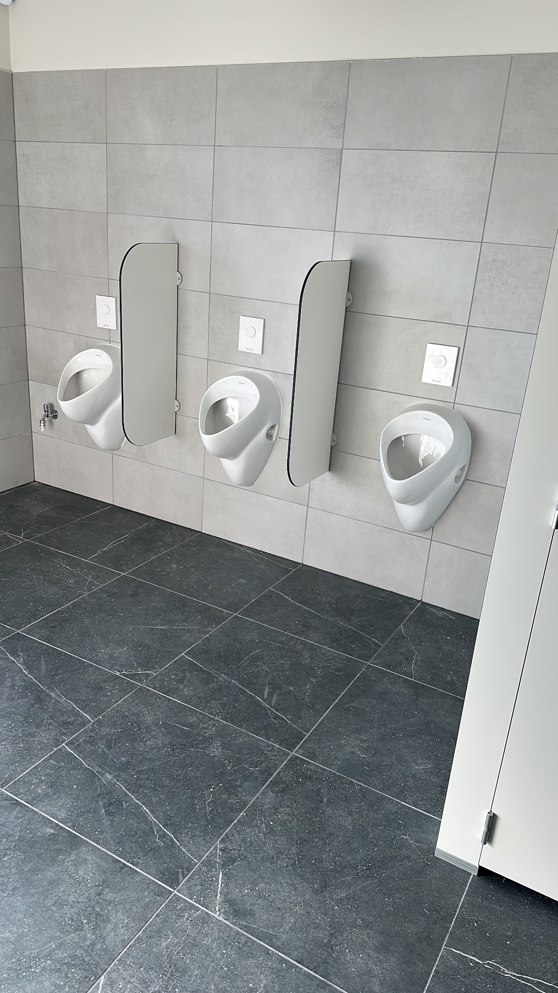 Nowoczesna toaleta publiczna z trzema białymi pisuarami i szarymi płytkami na ścianach i podłodze. Minimalistyczny design, czyste linie, oświetlenie górne.