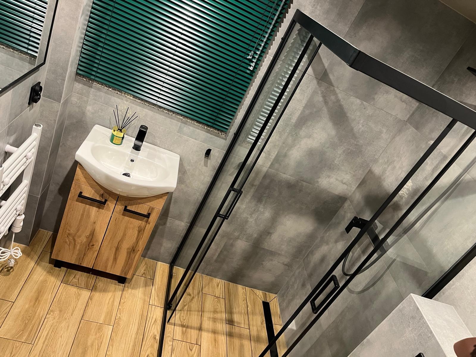 Nowoczesna łazienka z kabiną prysznicową typu walk-in, drewnianą szafką pod umywalką i zielonymi żaluzjami. Szare płytki na ścianach i podłodze imitują drewno.