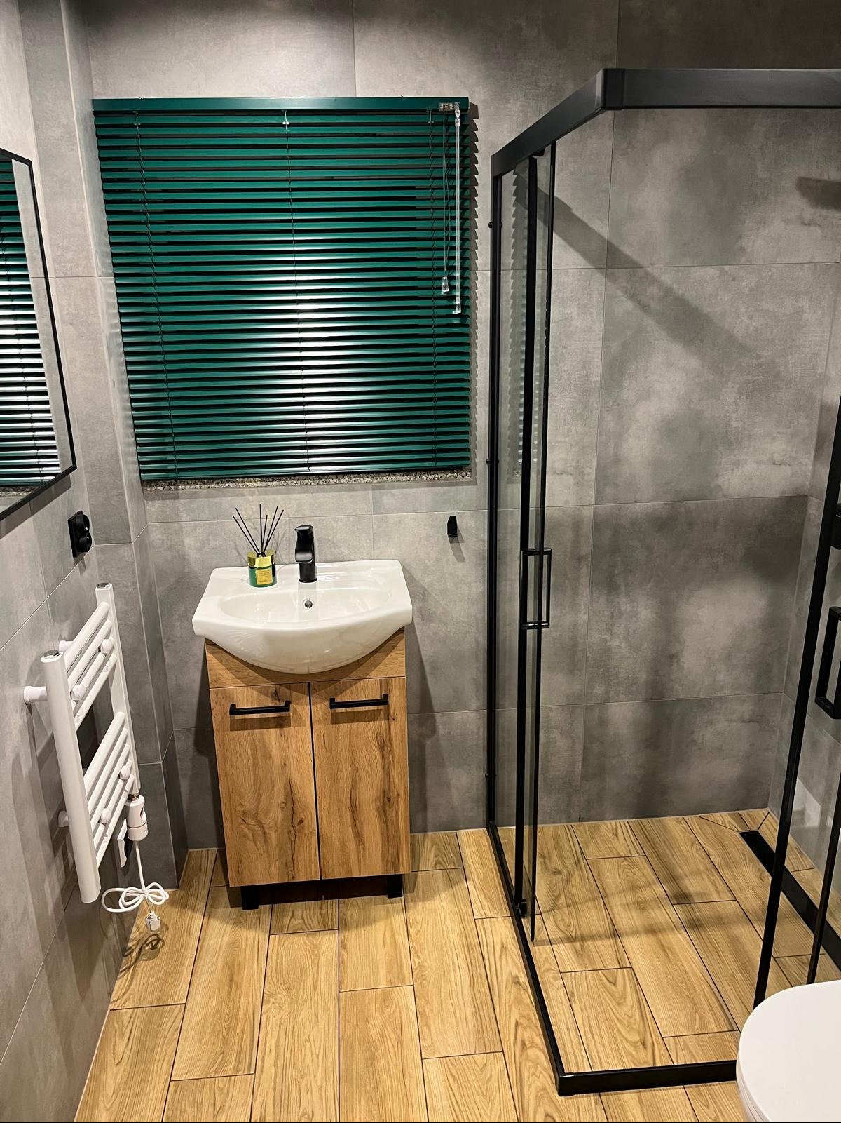Nowoczesna łazienka z szarą płytką, drewnianą szafką pod umywalką i kabiną prysznicową z czarnymi ramami. Akcent stanowi zielona roleta okienna.