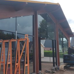 Mikołaj Jeliński Deerspaw - Montaż dużych okien fasadowych w budynku o konstrukcji drewnianej, widoczny pomarańczowy stojak na szyby i koparka w tle.