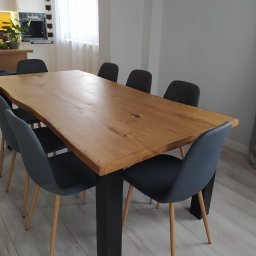 Mikołaj Jeliński Deerspaw - Stół z blatem z litego drewna o nieregularnych krawędziach, wsparty na czarnych, kwadratowych nogach, otoczony sześcioma krzesłami z szarą tapicerką i drewnianymi nogami, w jasnym wnętrzu.