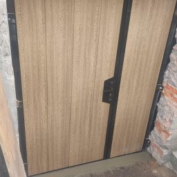Mikołaj Jeliński Deerspaw - Nowo zamontowana dwuskrzydłowa brama wjazdowa z panelami imitującymi drewno, osadzona w surowym otworze muru z widocznymi śladami zaprawy i gruzu.
