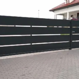 Nowoczesna, przesuwna brama wjazdowa w kolorze antracytu z poziomymi, metalowymi panelami i subtelnymi, srebrnymi akcentami, zamontowana przy posesji z białym domem i kostką brukową.