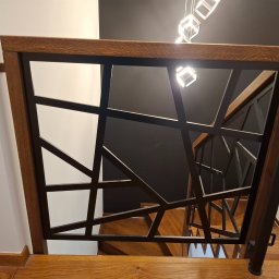 Mikołaj Jeliński Deerspaw - Nowoczesna balustrada schodowa z drewnianą ramą i czarnymi, geometrycznymi elementami metalowymi, widok z góry na drewniane schody i designerskie oświetlenie sufitowe.