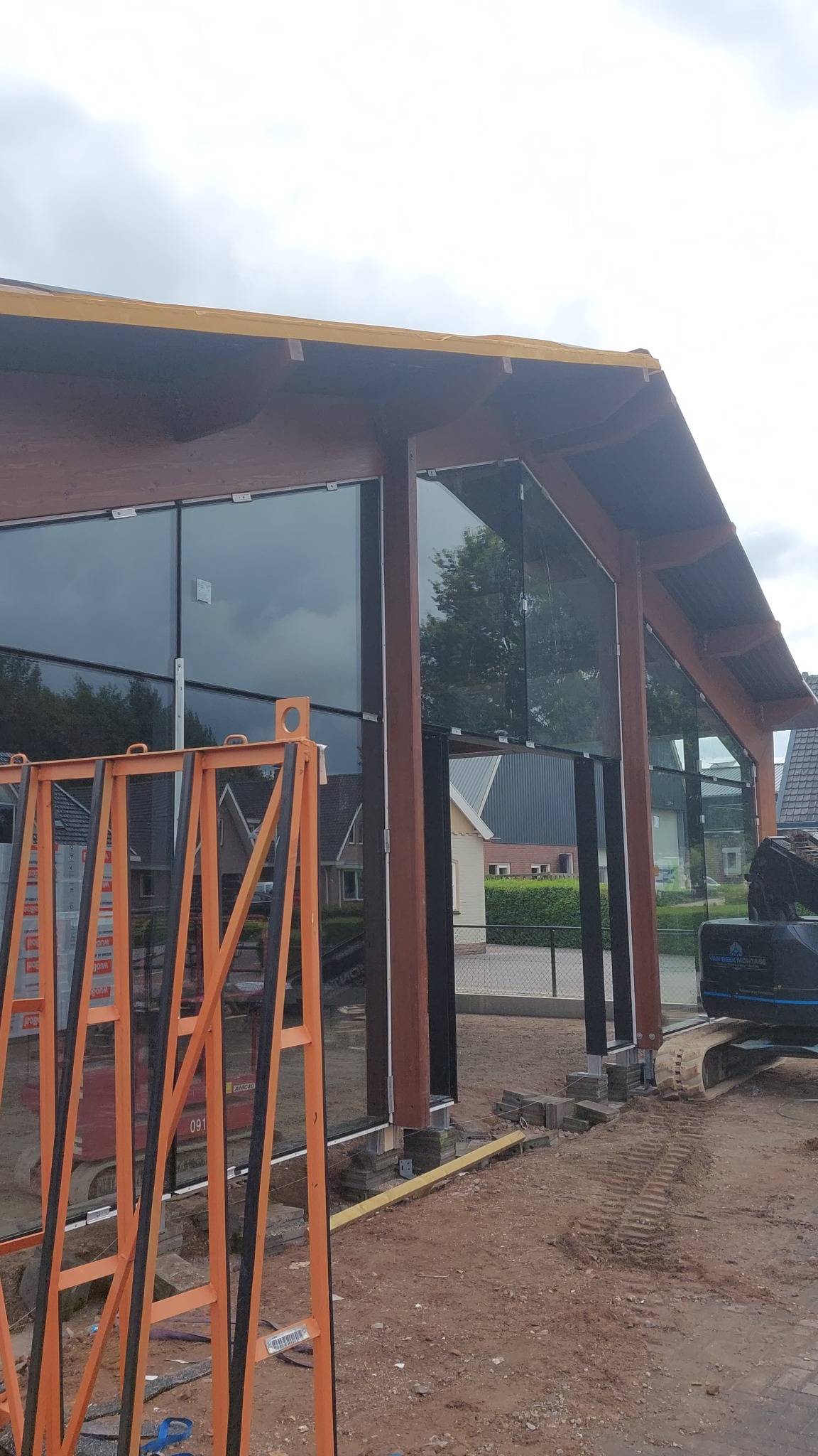 Montaż dużych okien fasadowych w budynku o konstrukcji drewnianej, widoczny pomarańczowy stojak na szyby i koparka w tle.