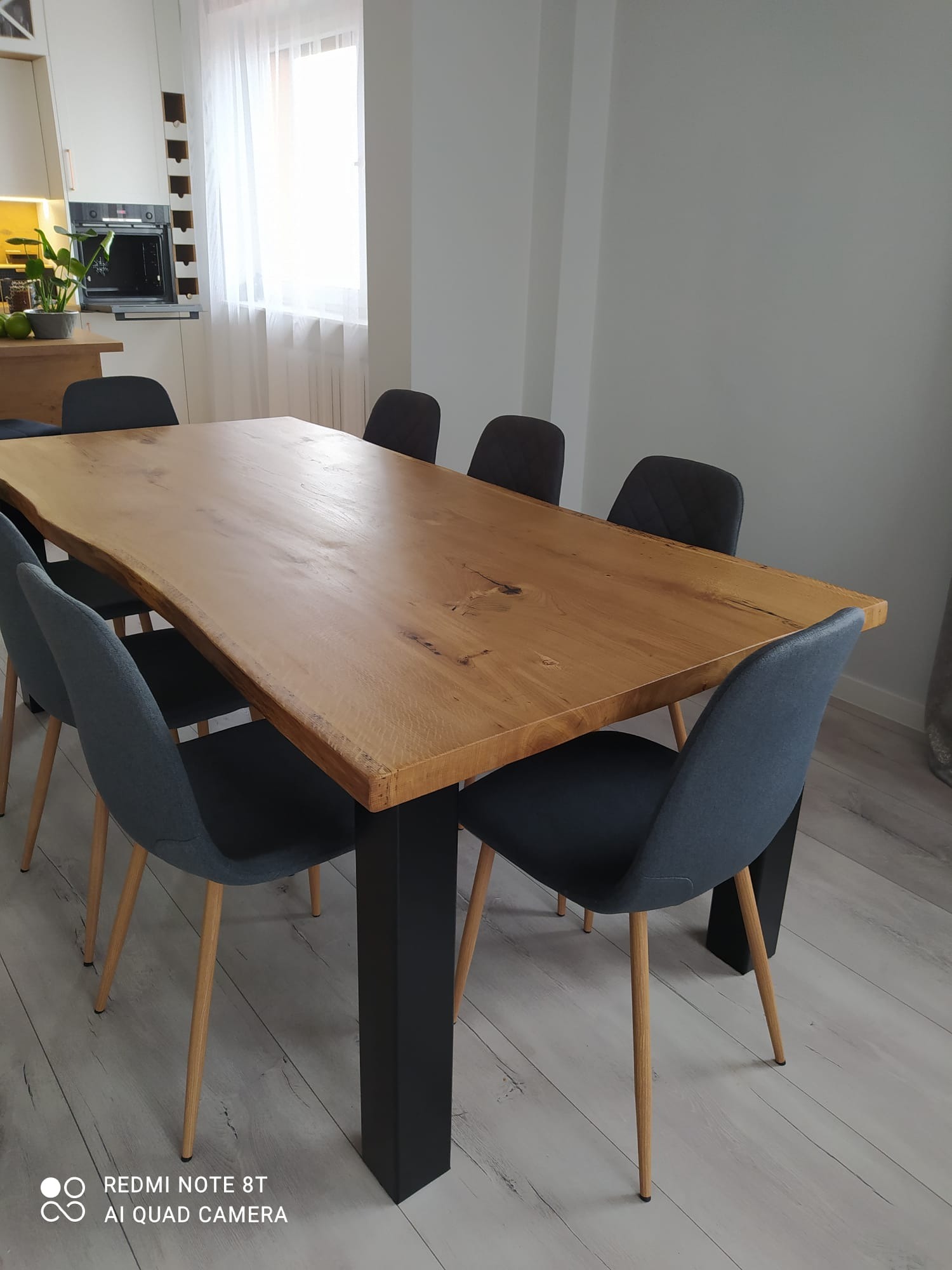 Stół z blatem z litego drewna o nieregularnych krawędziach, wsparty na czarnych, kwadratowych nogach, otoczony sześcioma krzesłami z szarą tapicerką i drewnianymi nogami, w jasnym wnętrzu.