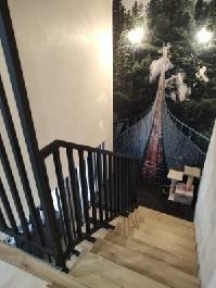 Klatka schodowa z drewnianymi stopniami i czarną, metalową balustradą. Na ścianie fototapeta z motywem leśnym i mostem wiszącym.