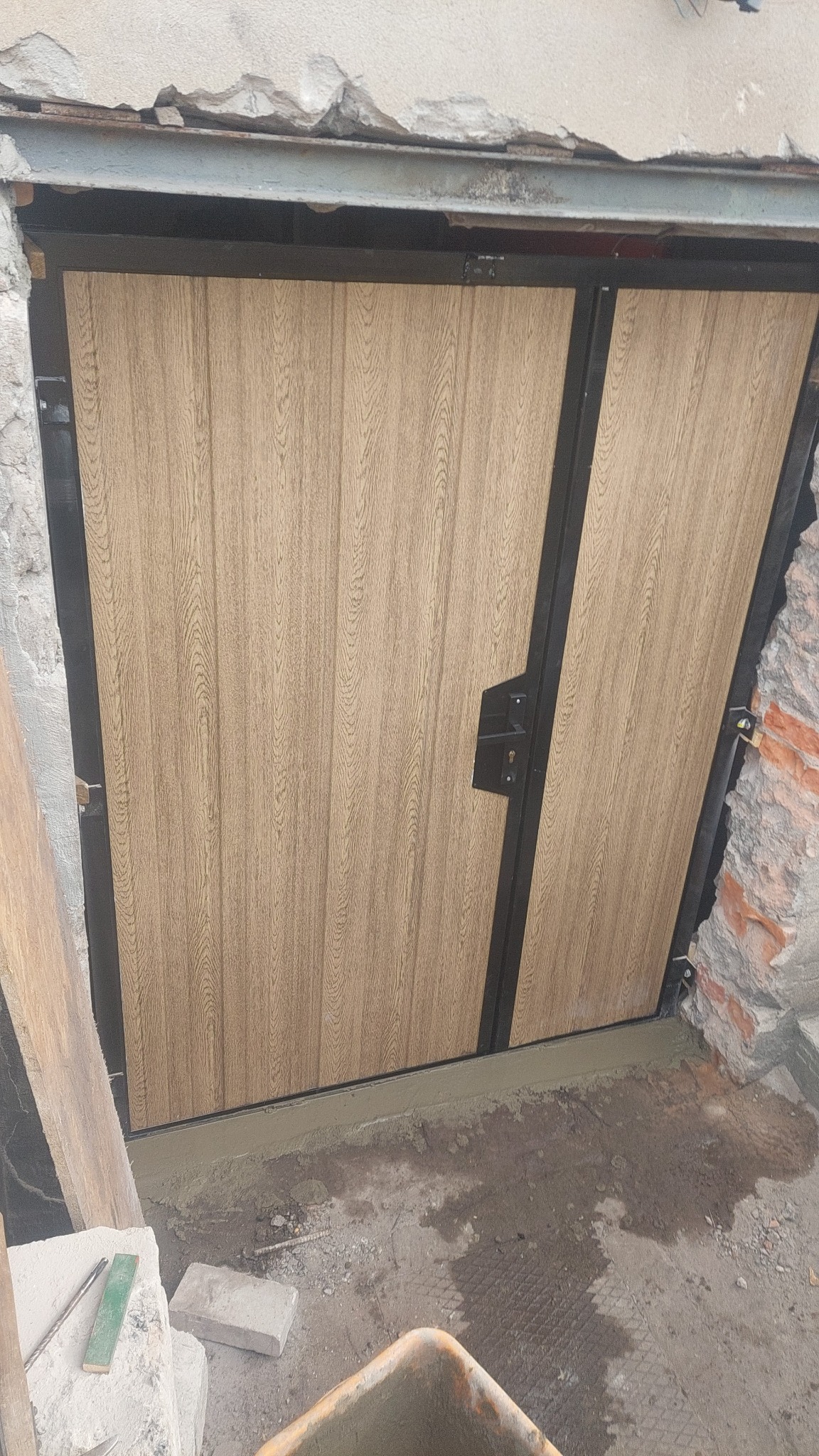 Nowo zamontowana dwuskrzydłowa brama wjazdowa z panelami imitującymi drewno, osadzona w surowym otworze muru z widocznymi śladami zaprawy i gruzu.