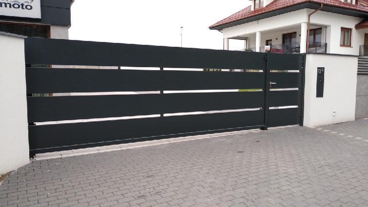 Nowoczesna, przesuwna brama wjazdowa w kolorze antracytu z poziomymi, metalowymi panelami i subtelnymi, srebrnymi akcentami, zamontowana przy posesji z białym domem i kostką brukową.
