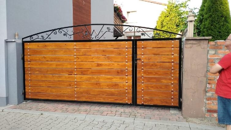 Dwuskrzydłowa brama wjazdowa z poziomych desek w ciepłym odcieniu brązu, osadzona w czarnej metalowej ramie z ozdobnym, kutym elementem w górnej części, widoczna od strony posesji.