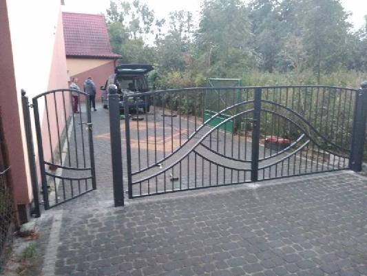 Otwarta, czarna brama wjazdowa z dekoracyjnym, falistym wzorem, widoczna z zewnątrz posesji, z dwoma osobami i zaparkowanym samochodem w tle.