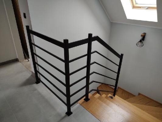 Nowoczesna, czarna balustrada schodowa z poziomymi prętami, zamontowana przy drewnianych schodach w jasnym wnętrzu z oknem dachowym i designerską lampą na ścianie.