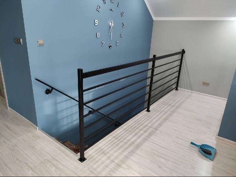 Czarna, pozioma balustrada z metalowych prętów na antresoli, ściana pomalowana na niebiesko, na ścianie zegar, na podłodze szczotka do zamiatania.