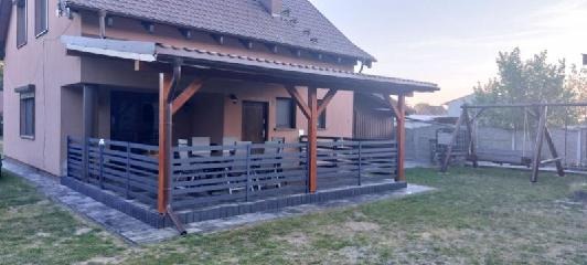 Zadaszony taras z drewnianą konstrukcją i szarą, poziomą balustradą, otoczony kostką brukową, z widocznym huśtawką ogrodową i trawnikiem.