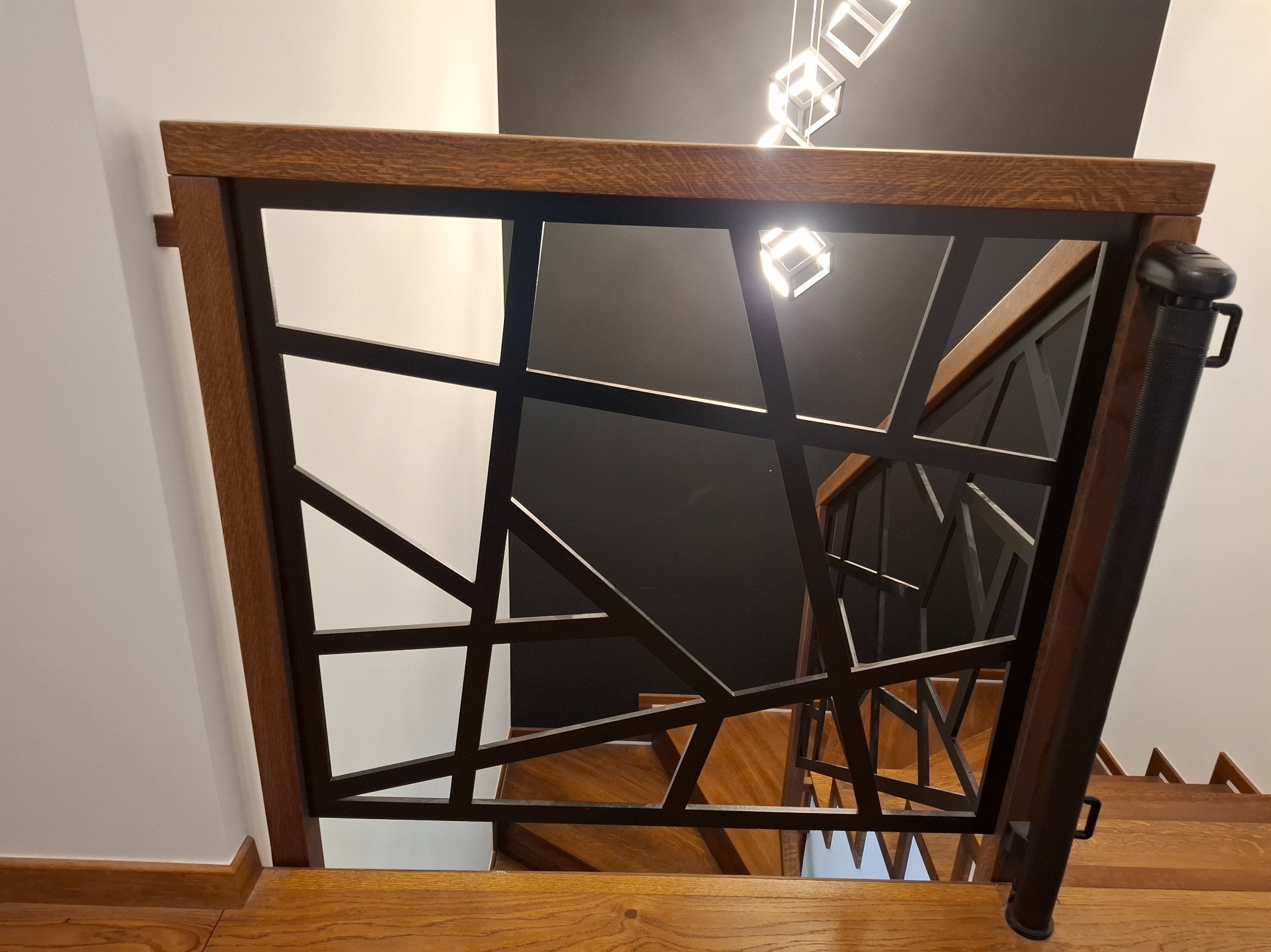 Nowoczesna balustrada schodowa z drewnianą ramą i czarnymi, geometrycznymi elementami metalowymi, widok z góry na drewniane schody i designerskie oświetlenie sufitowe.