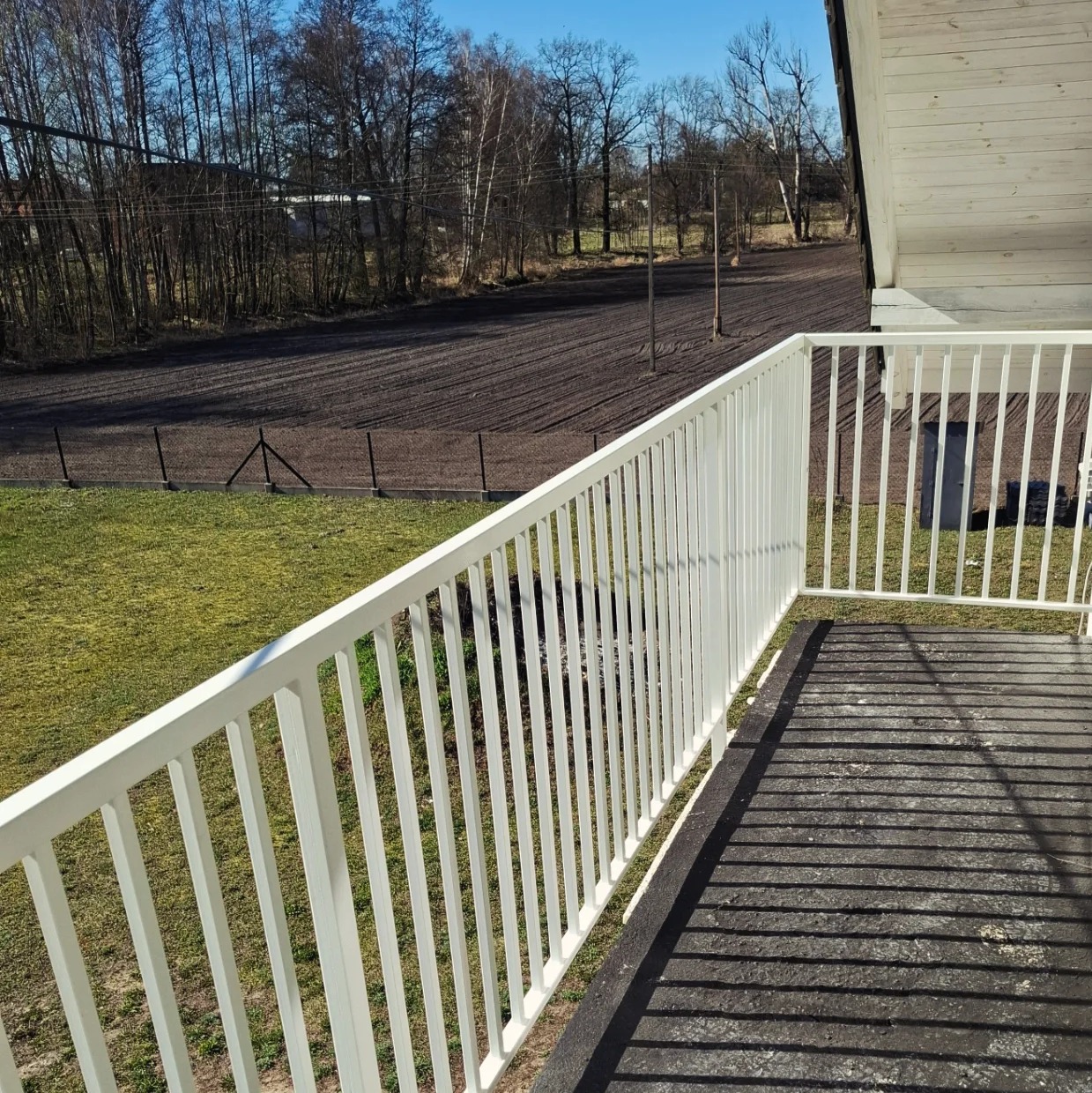 Biała balustrada balkonowa z widokiem na zaorane pole i drzewa w tle, w słoneczny dzień.