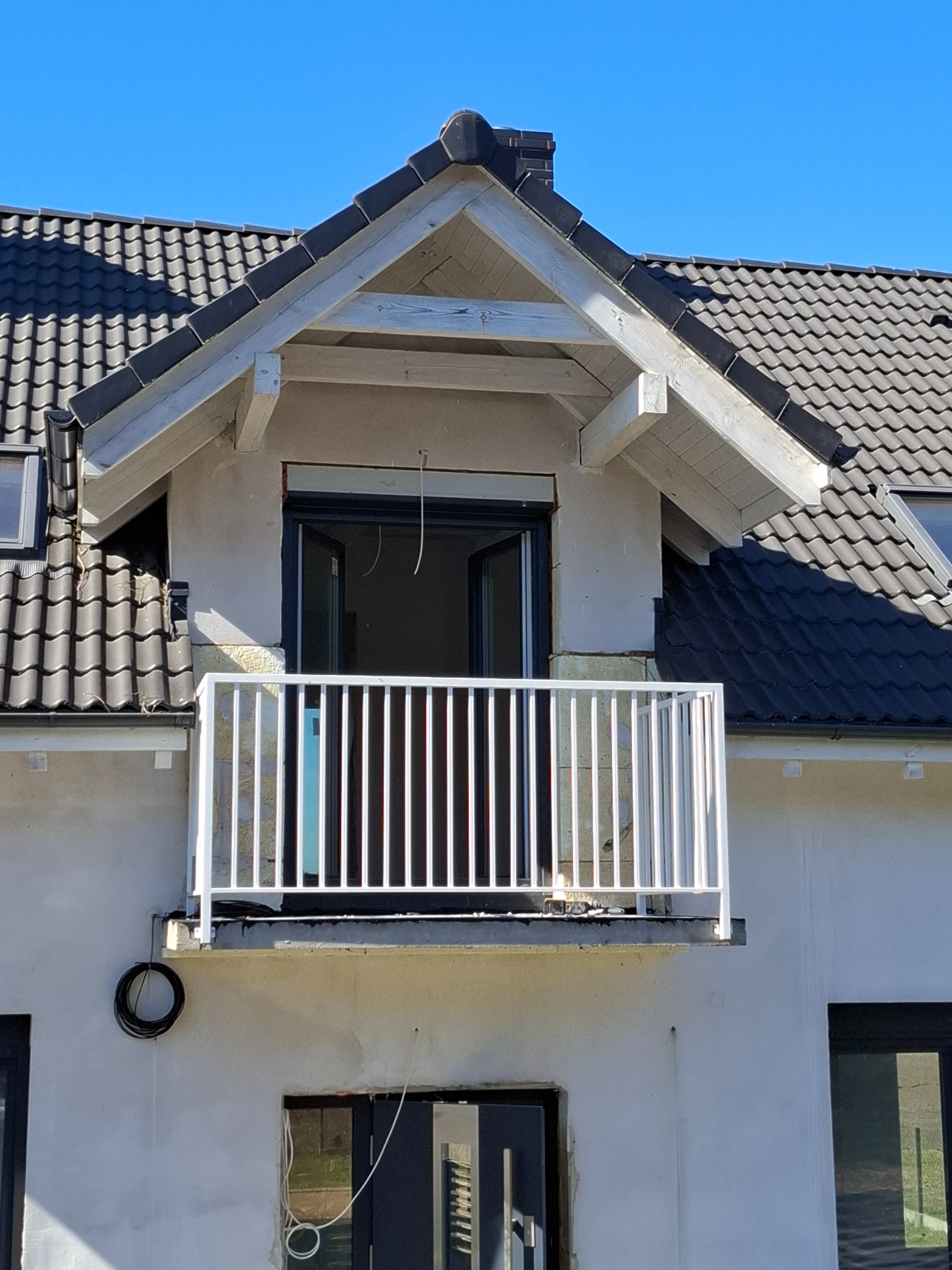 Balkon z białą, pionową balustradą na tle jasnej elewacji domu z ciemną dachówką i otwartymi drzwiami balkonowymi, widoczne okablowanie na ścianie.