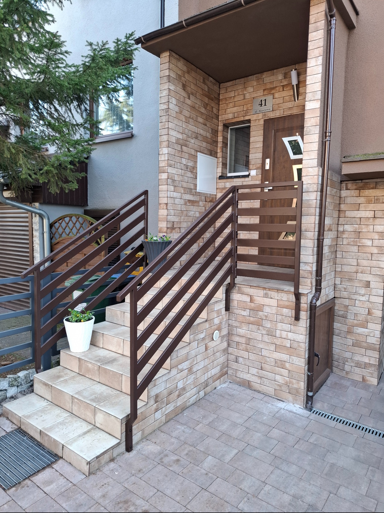 Zewnętrzne schody wejściowe do domu z brązową, poziomą balustradą, doniczkami z kwiatami i elewacją z cegły.