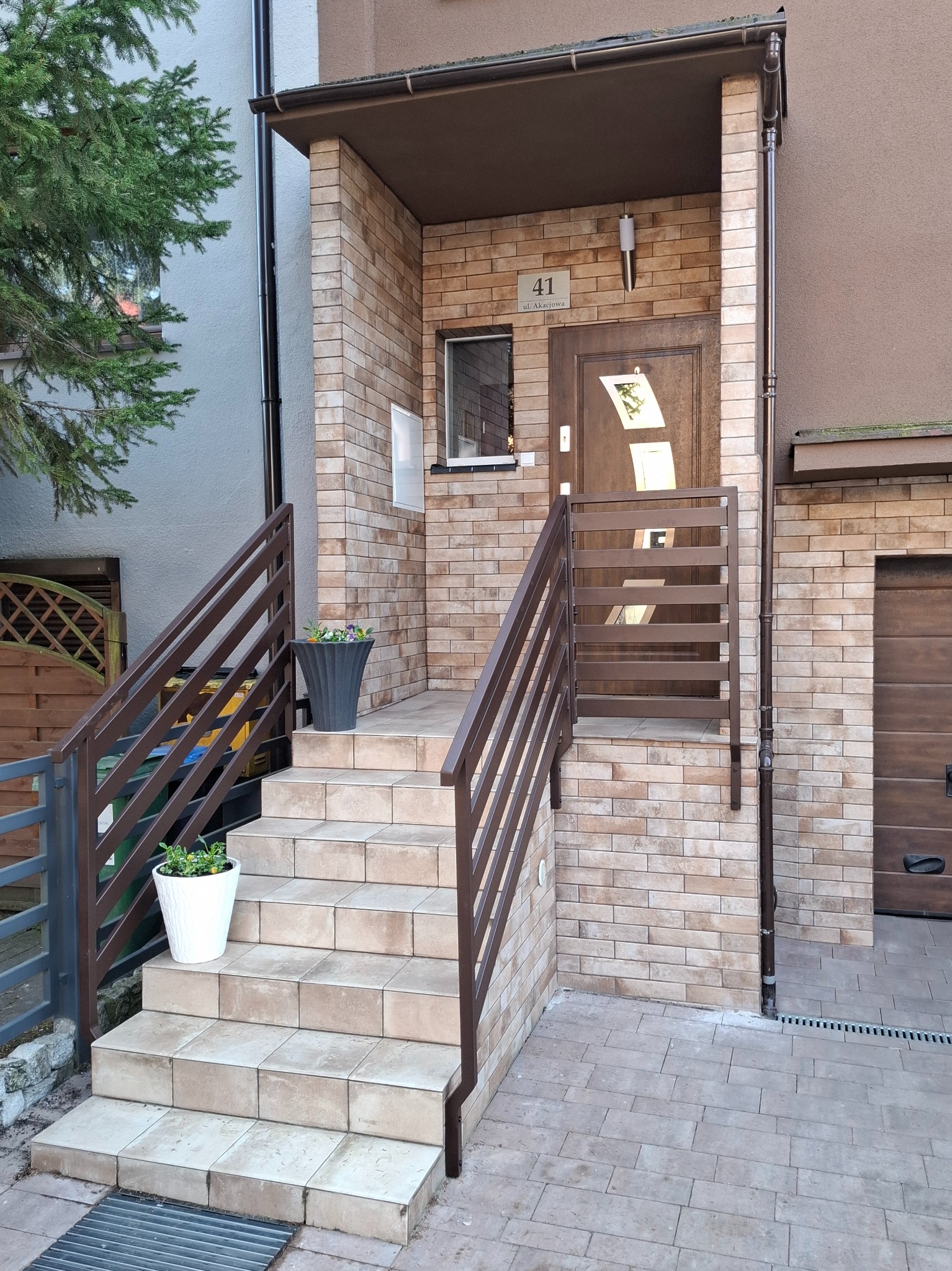 Wejście do domu z brązowymi schodami i balustradami z poziomych profili, donice z kwiatami po bokach, elewacja z cegły klinkierowej.