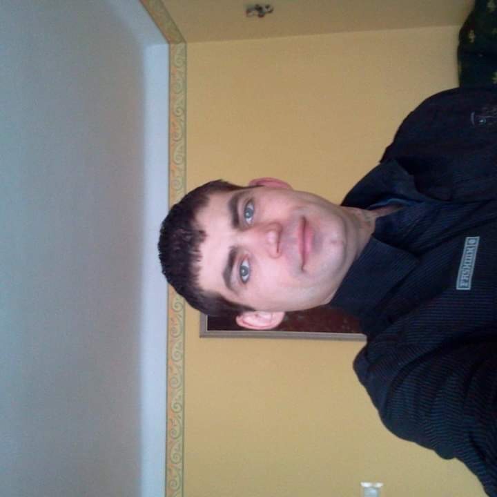 Portret mężczyzny z krótkimi włosami na tle ściany z żółtą farbą i dekoracyjnym paskiem przy suficie.