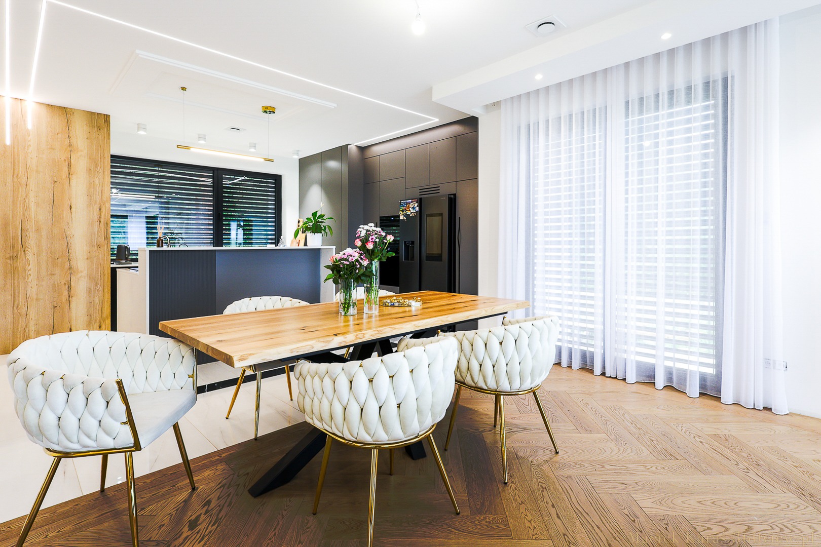 Nowoczesna jadalnia z drewnianym stołem o nieregularnych krawędziach, białymi krzesłami z plecionym oparciem na złotych nóżkach, parkietem w jodełkę i oświetleniem LED.