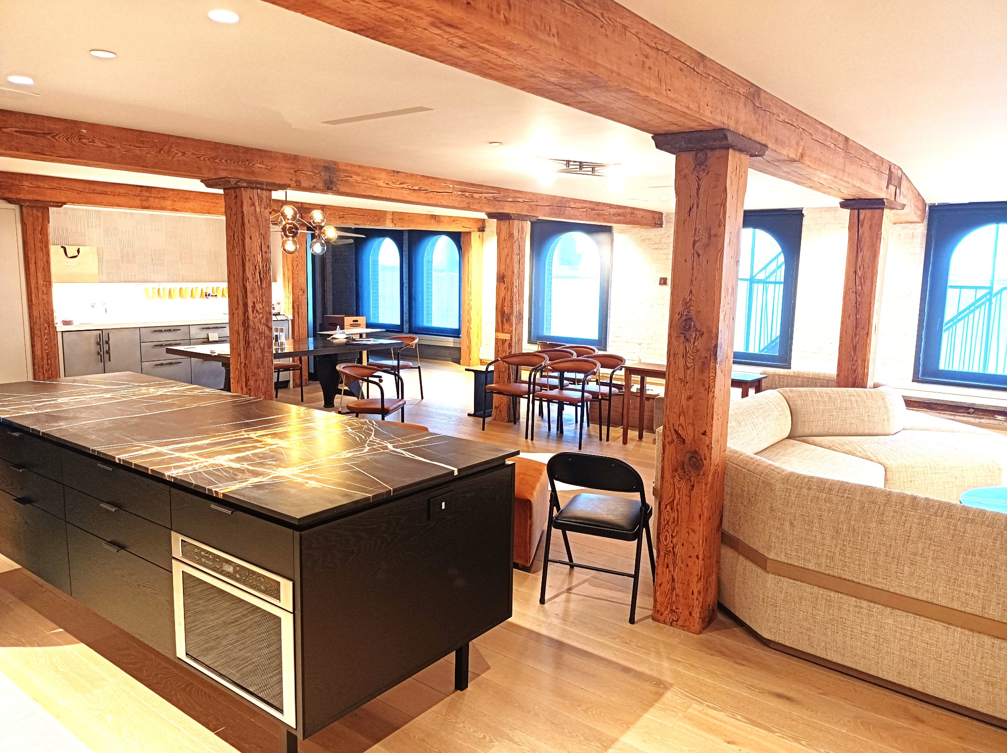 Przestronne wnętrze loftu z drewnianymi belkami stropowymi, wyspą kuchenną z blatem z efektem marmuru, dużymi oknami w stylu industrialnym i narożną sofą w neutralnych barwach.