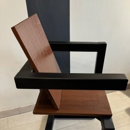 DCwelding - Krzesło z drewnianym siedziskiem i oparciem w odcieniach brązu, z czarną, metalową ramą na tle ściany w biało-grafitowe pasy.