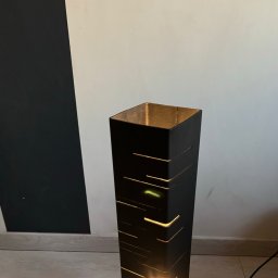 DCwelding - Czarna, metalowa lampa podłogowa o geometrycznym wzorze, z wyciętymi paskami przepuszczającymi ciepłe światło, na tle dwukolorowej ściany.