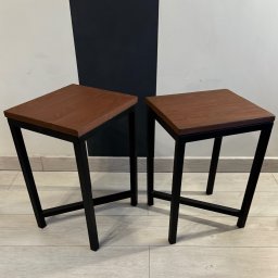 DCwelding - Dwa identyczne stołki barowe z kwadratowymi siedziskami z ciemnego drewna i czarnymi metalowymi nogami o geometrycznym kształcie na tle ściany w biało-czarne pasy.
