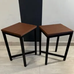 Dwa identyczne stołki barowe z kwadratowymi siedziskami z ciemnego drewna i czarnymi metalowymi nogami o geometrycznym kształcie na tle ściany w biało-czarne pasy.