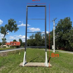 DCwelding - Wysoka, metalowa konstrukcja ramy, prawdopodobnie pod billboard lub reklamę, z widocznymi wymiarami 2m x 8m, umieszczona na betonowej podstawie na trawiastym terenie, z budynkami i słupem...