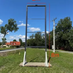 Wysoka, metalowa konstrukcja ramy, prawdopodobnie pod billboard lub reklamę, z widocznymi wymiarami 2m x 8m, umieszczona na betonowej podstawie na trawiastym terenie, z budynkami i słupem...