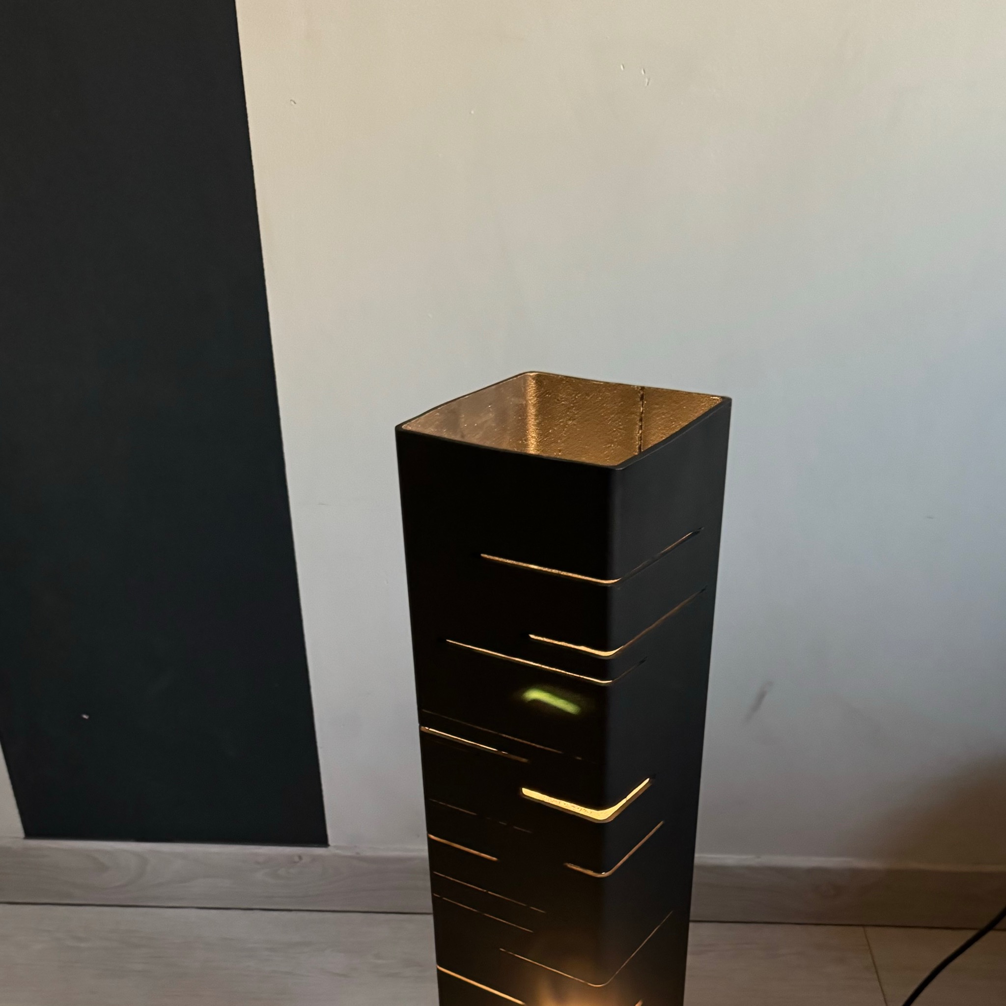 Czarna, metalowa lampa podłogowa o geometrycznym wzorze, z wyciętymi paskami przepuszczającymi ciepłe światło, na tle dwukolorowej ściany.